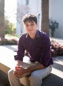 Erubey sits on stone bench with flowers, trees, and building behind him wearing a purple button down with sleeves rolled up and khaki pants.
