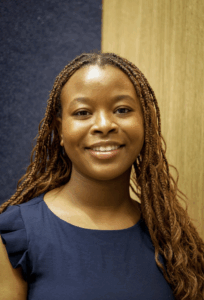 Isis is in front of a corner where blue and wood paneled wall meet wearing a blue shirt and hair in braids.