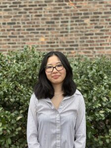 Ana in front of bushes and brick wall wearing her hair down, glasses, and white striped button dowin shirt.