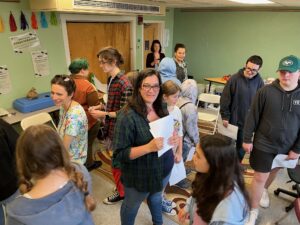 Adult staff person wearing glasses and holding papers looks at camera while class moves around.